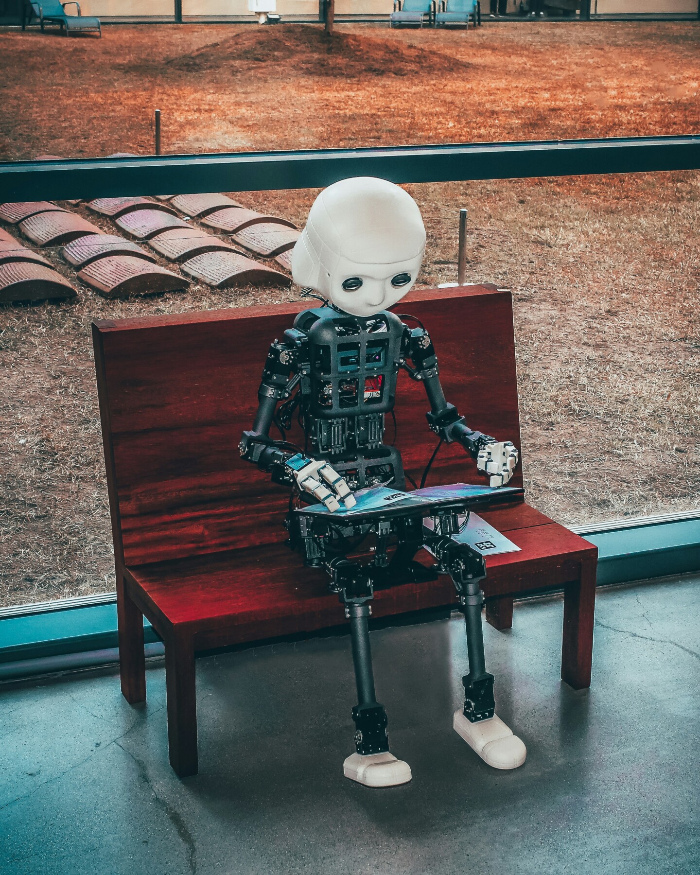 Ein humanoider Roboter mit weißem Kopf und schwarzen mechanischen Gliedmaßen sitzt auf einer Holzbank und betrachtet ein Tablet. Im Hintergrund befindet sich ein brauner Rasen.