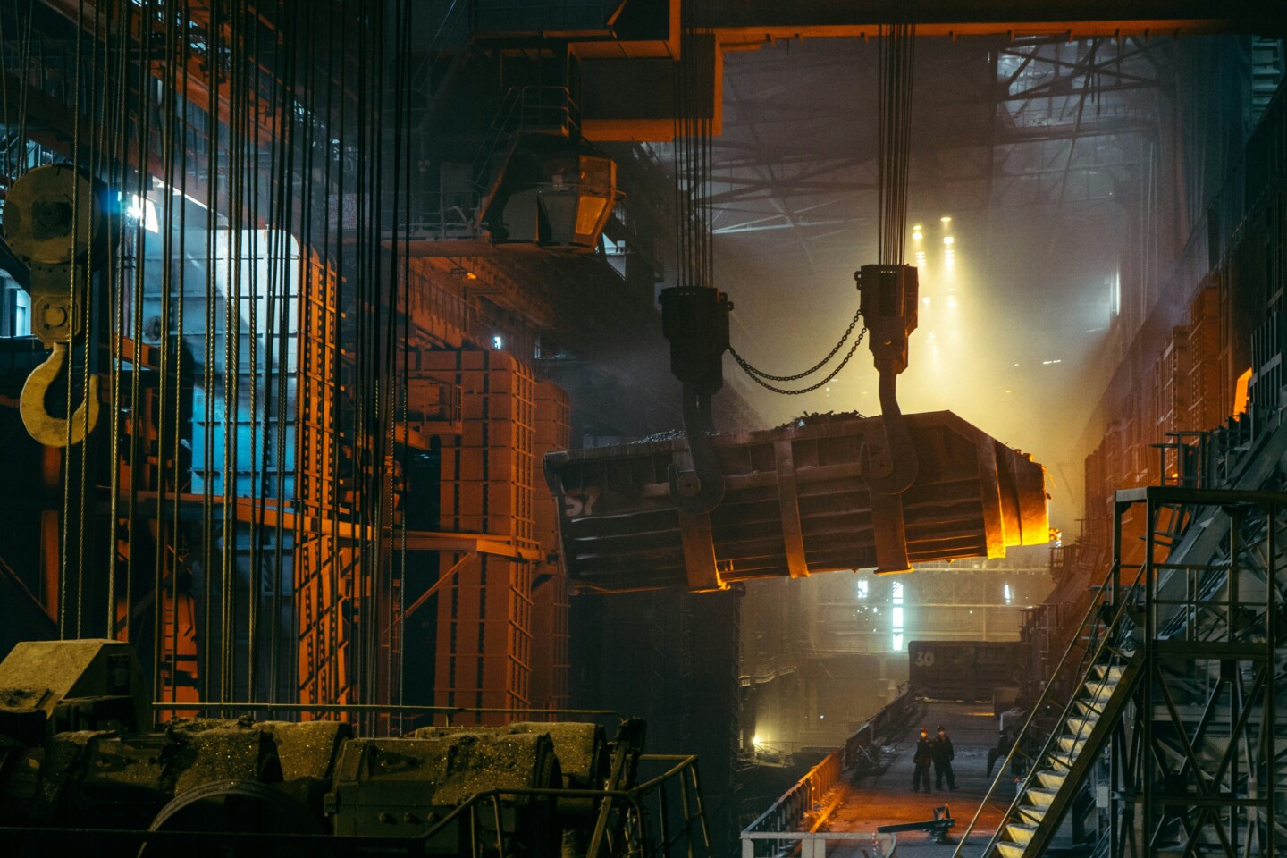 Innenansicht eines Industrie-Stahlwerks mit Haken und schweren Maschinen, die metallisches Material bewegen. Arbeiter sind im Hintergrund sichtbar sowie die Arbeitsplaztzbeleuchtung. Die Szene ist von warmem, gelblichem Licht durchdrungen.