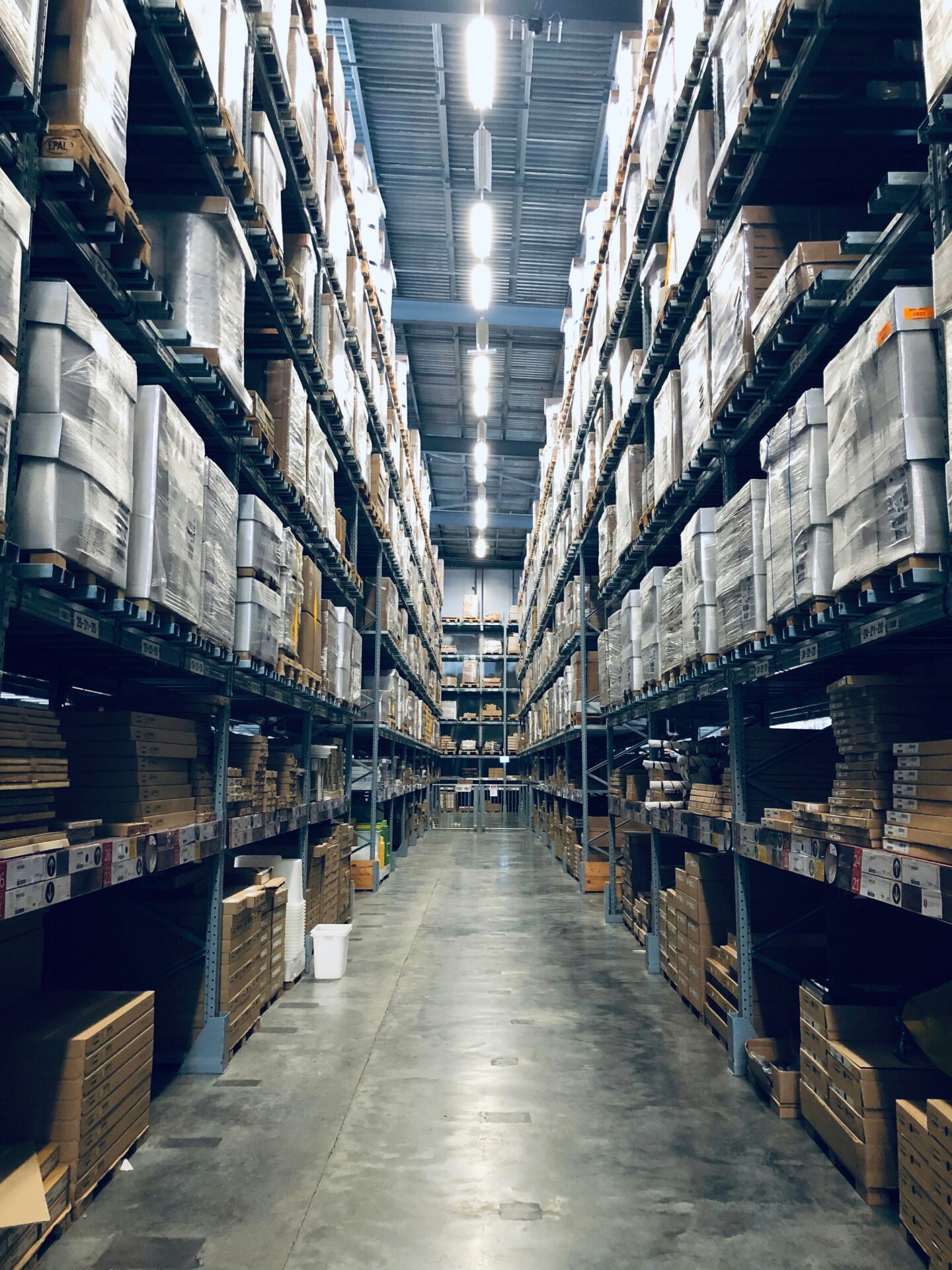 Ein langer Gang in einem Lagerhaus, umgeben von hohen Regalen, die mit Kartons und verpackten Waren gefüllt sind. T5/T8 Beleuchtung