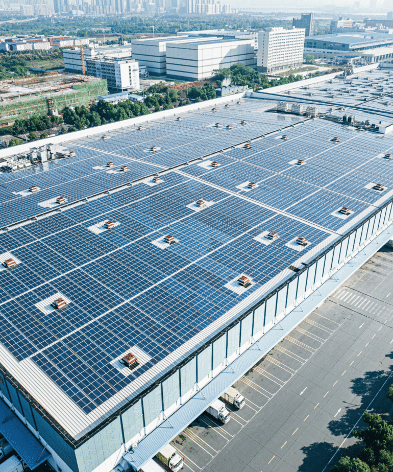 Luftaufnahme eines Fabrikdachs mit Solar Panelen. Im Hintergrund sieht man die Skyline einer Stand mit Hochhäusern.